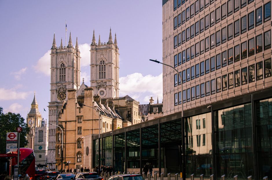 The image shows the exterior of Westminster Abbey with its Gothic architecture, featuring two tall, intricately detailed towers topped with spires and decorative finials. The facade is constructed fro