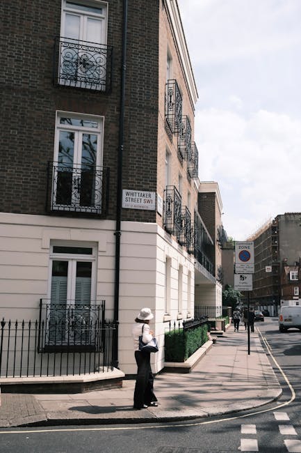 A woman wearing a wide-brimmed hat, white shirt, and dark trousers stands on the sidewalk outside a multi-storey residential building on Whittaker Street SW1. The building has a brick facade with mult